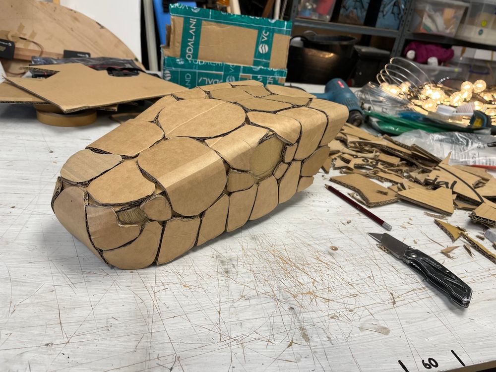 The previous cardboard snake head has now been covered in large cardboard scales that meet with thin gaps between them to create a pattern of lines over the surface The scales wrap around the edges softening the original boxy look. Gaps have been left where the eyes and nostrils are. As before, the head lies on the workbench. Next to it is piles of little cardboard scraps, a box knife and a pencil.