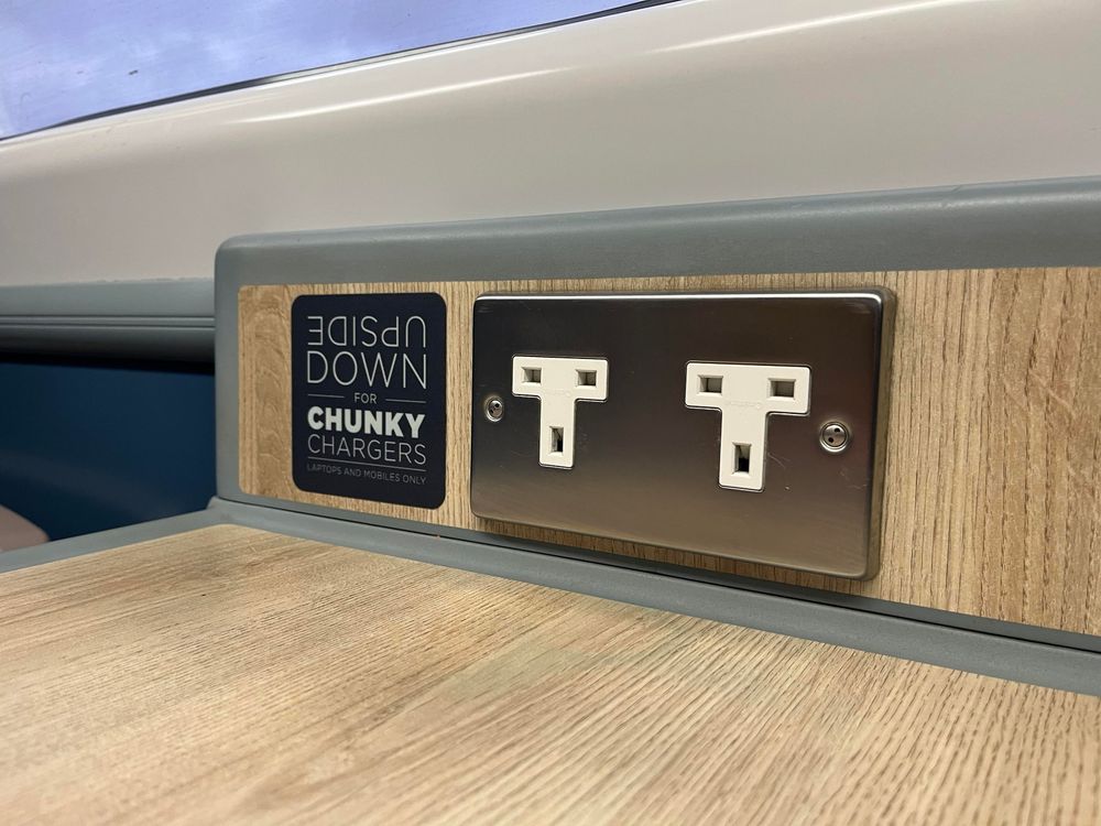 A pair of UK power sockets under a window just above the train table that have been inserted upside down. Next to it is a small sign that reads, “Upside down for chunky chargers”