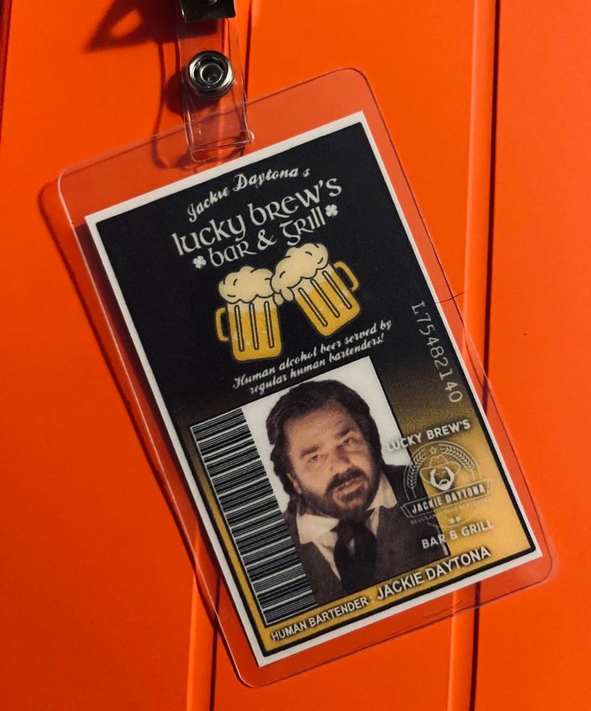 An ID badge sitting on an orange iPad case. The laminated card purports to be for "Human Bartender Jackie Daytona" and features a barcode alongside a photograph of actor Matt Berry in his role in the comedy series "What We Do in the Shadows". The ID states that it is from "Jackie Daytona's Lucky Brew's Bar & Grill".