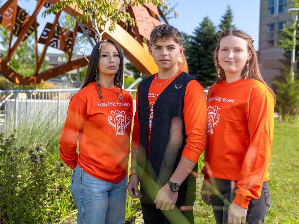 Three students stand outside the Wampum Learning Lodge wearing orange shirts that read, "Every Child Matters".