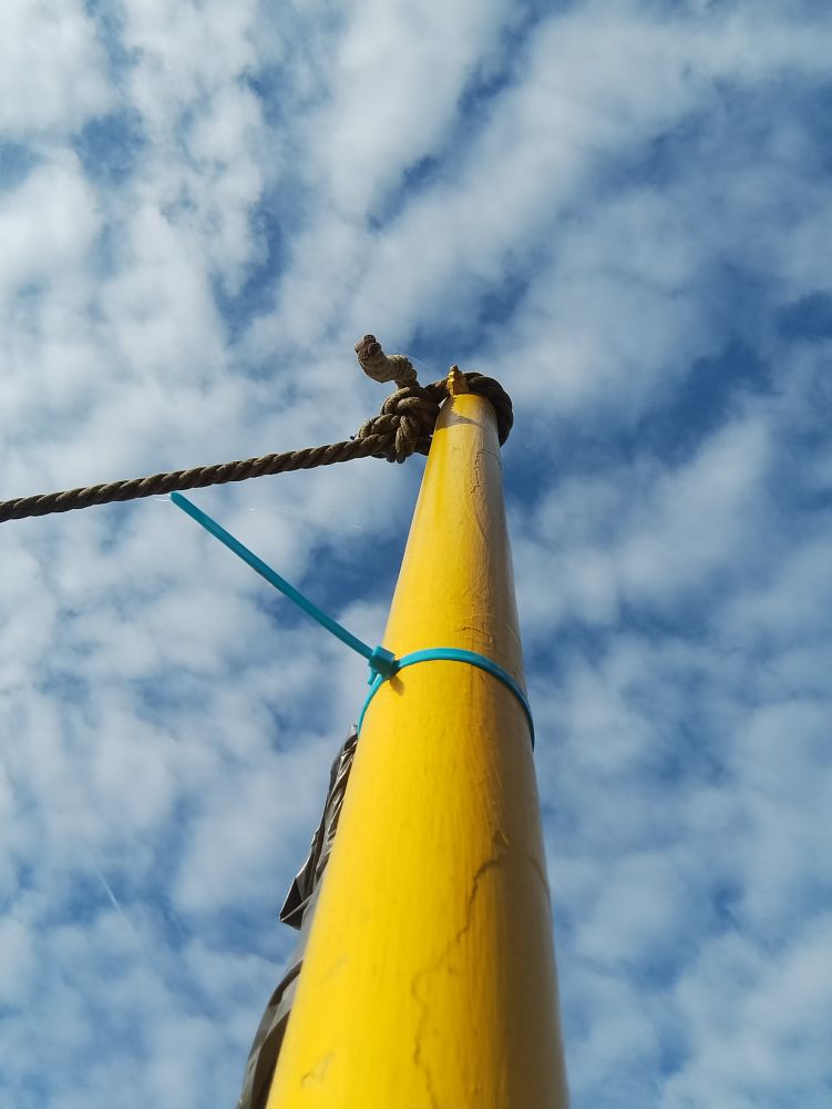photo. a yellow metal post, with a rope at the top, a cable binder round the middle, a piece of plastic is barely visible behind the post. photographed from down below so that it ascends into a blue and cloudy sky 