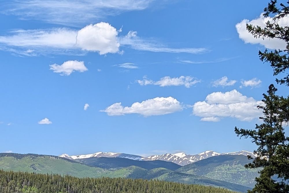 Green hills with pine trees and snow covered mountains in the background with blue skies and a few fluffy clouds above. 