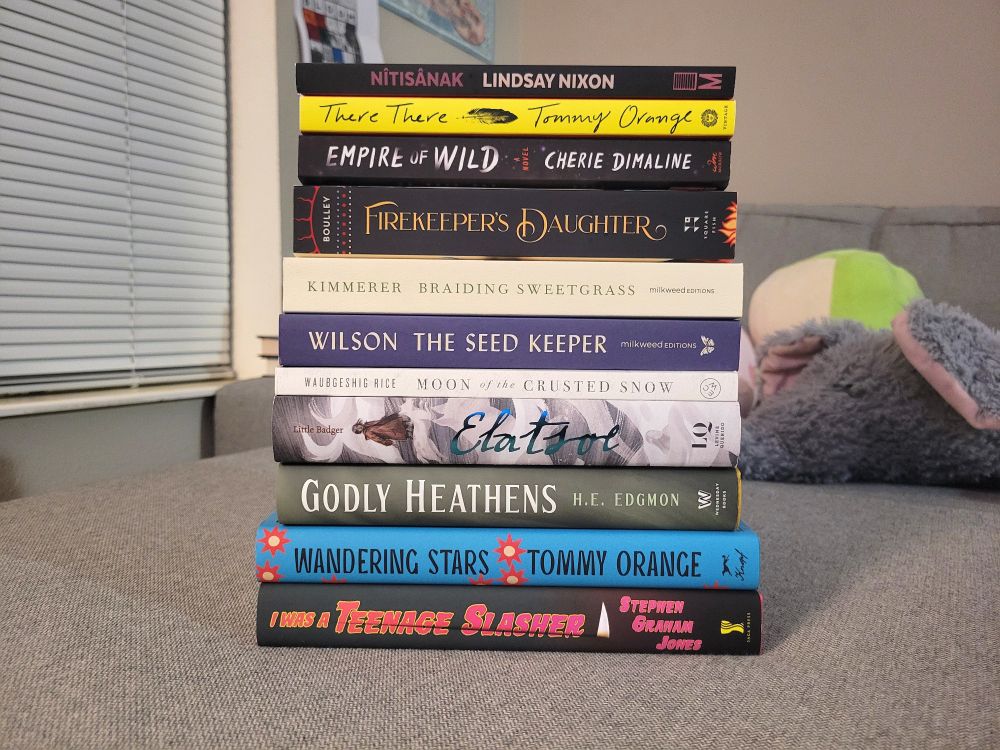 There's a stack of books written by indigenous authors during on top of a grey couch in the picture. The title are as followed from bottom to top of the book pile:
I was a teenage slasher by Stephen Graham Jones 
Wandering stars by Tommy Orange 
Godly heathens by H.E. Edgmon 
Elatsoe by Darcy Little Badger
Moon of the crusted snow by Waubgeshig Rice
The Seed Keeper by Diane Wilson 
Braiding sweetgrass by Robin Wall kimmerer 
The firekeeper's daughter by Angeline Boulley
Empire of Wild by Cherie Dimaline 
There, There by Tommy Orange
Nîtîsanak by Jas M. Morgan