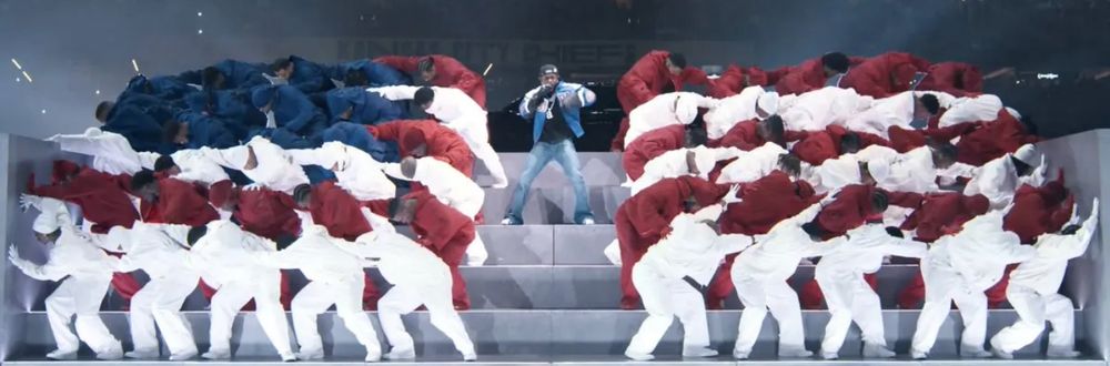 Kendrick Lamar performing at the Super Bowl half time show, standing in the center of an all Black groups of dancers in red, white, and blue, arranged like an American flag that is splitting in the middle.