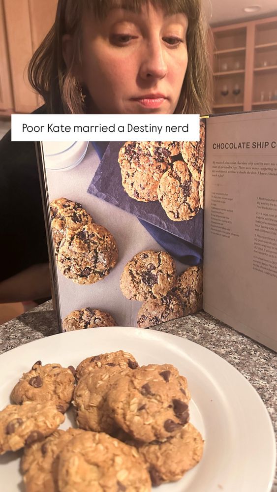 Kate peering over a Destiny 2 cookbook, at her batch of “chocolate shop cookies”, a batch she made for Joechuck’s bday. Oatmeal, chocolate chip situation on a clean white plate 