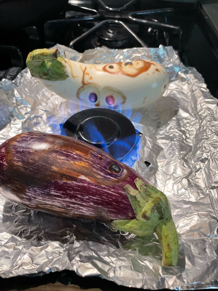Two heirloom eggplant on a sheet of foil around an open gas flame. The flame is burning shapes into the eggplant skin and they look like flaming cartoon eyes