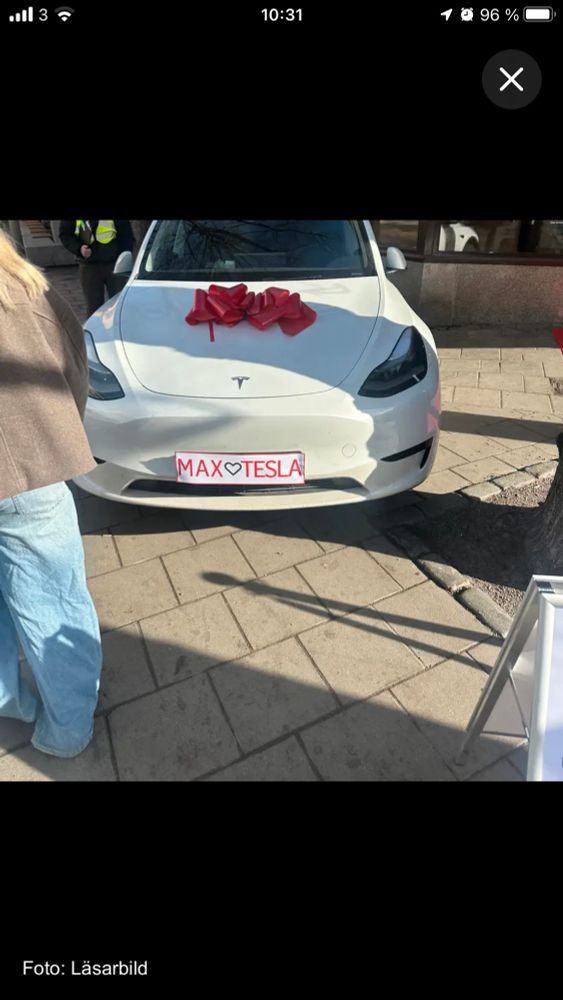Image of a white Tesla with a license plate “Max ❤️ Tesla” and red ribbons on the hood.