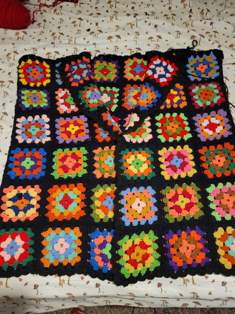 A crochet multicolored granny square blanket laid on a bed in the shape of a sleeveless jacket with lapels
