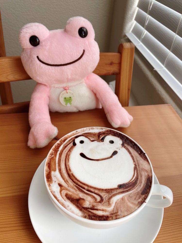 a pink plush frog sits at a wooden table smiling. a hot chocolate sits in front of him on the table in a white mug and saucer. the hot chocolate has a frog face drawn in the milk