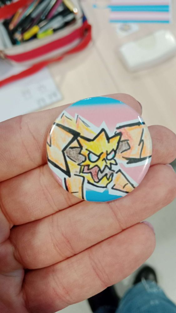 Badge supporting a cartoonish rey dau on front of the trans pride flag