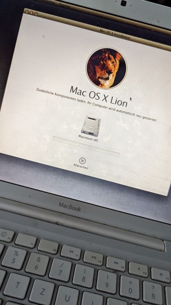 Ein altes, weißes MacBook Pro mit dem Installationsbildschirm. Zu sehen ist der kreisförmig ausgeschnittene Kopf eines Löwen. Darunter steht in dunkelgrauer Schrift auf weißem Grund: "Mac OS X Lion. Zusätzliche Komponenten laden. Ihr Computer wird automatisch neu gestartet". Der Fortschrittsbalken steht bei 0%. Dem MacBook sieht man an, dass es stark gebraucht und alt ist.