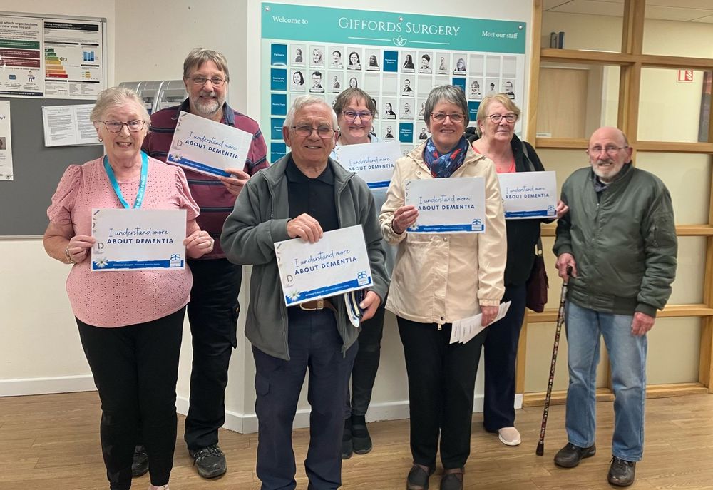 Group of six men and women smiling and holding 'I understand more about dementia' signs. Pictured indoors in a GP surgery premises.