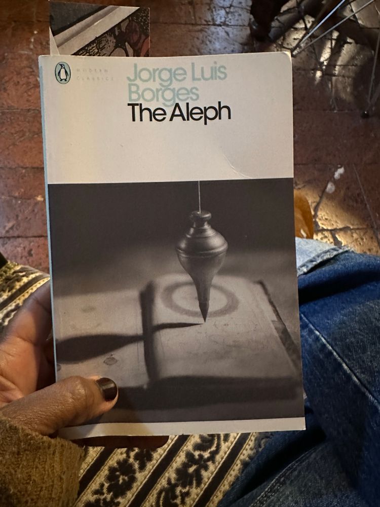 Book cover with the name Jose Luis Borges and title The Aleph on the front. A picture of a pendulum is below. A hand holds the book. 