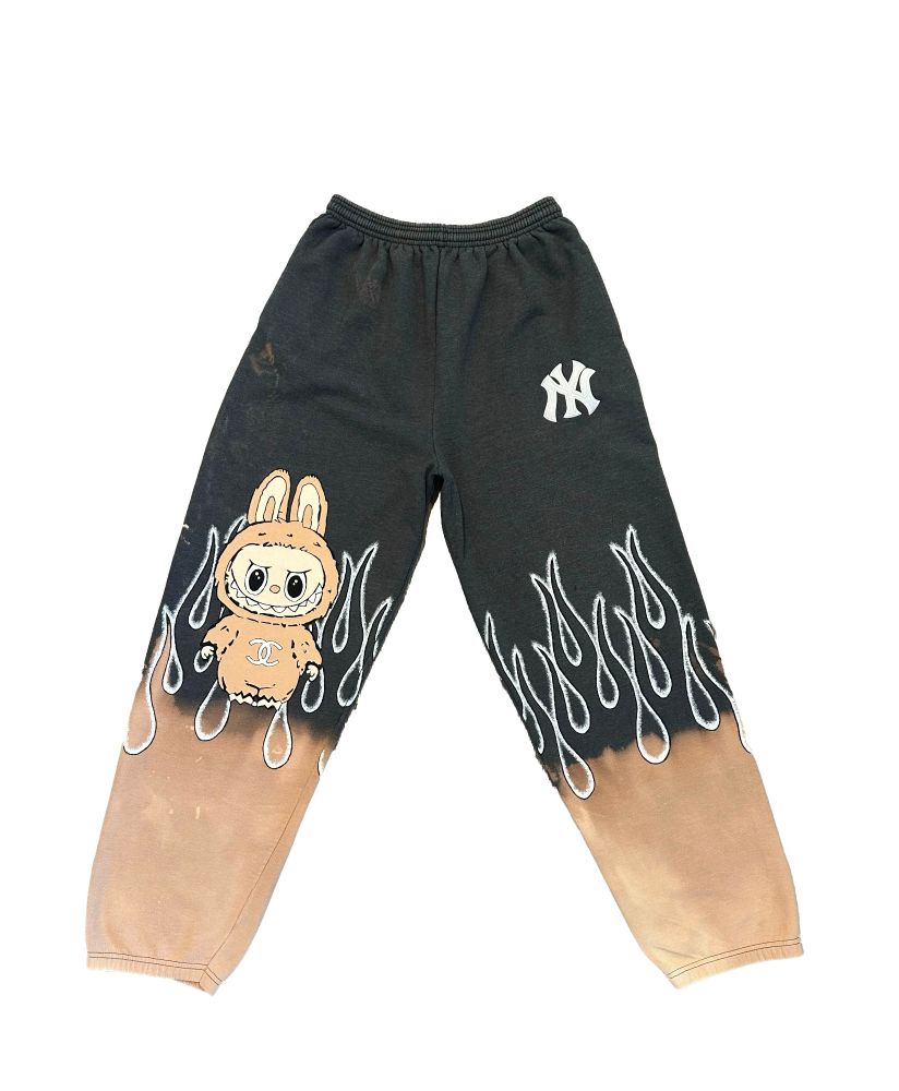A pair of sweat pants/joggers. They are black with bleaching at the ankles to the mid calf, and white painted flames over the top of the bleach to the lower thigh area. On the right leg is a beige labubu figure, on the left is a NY emblem