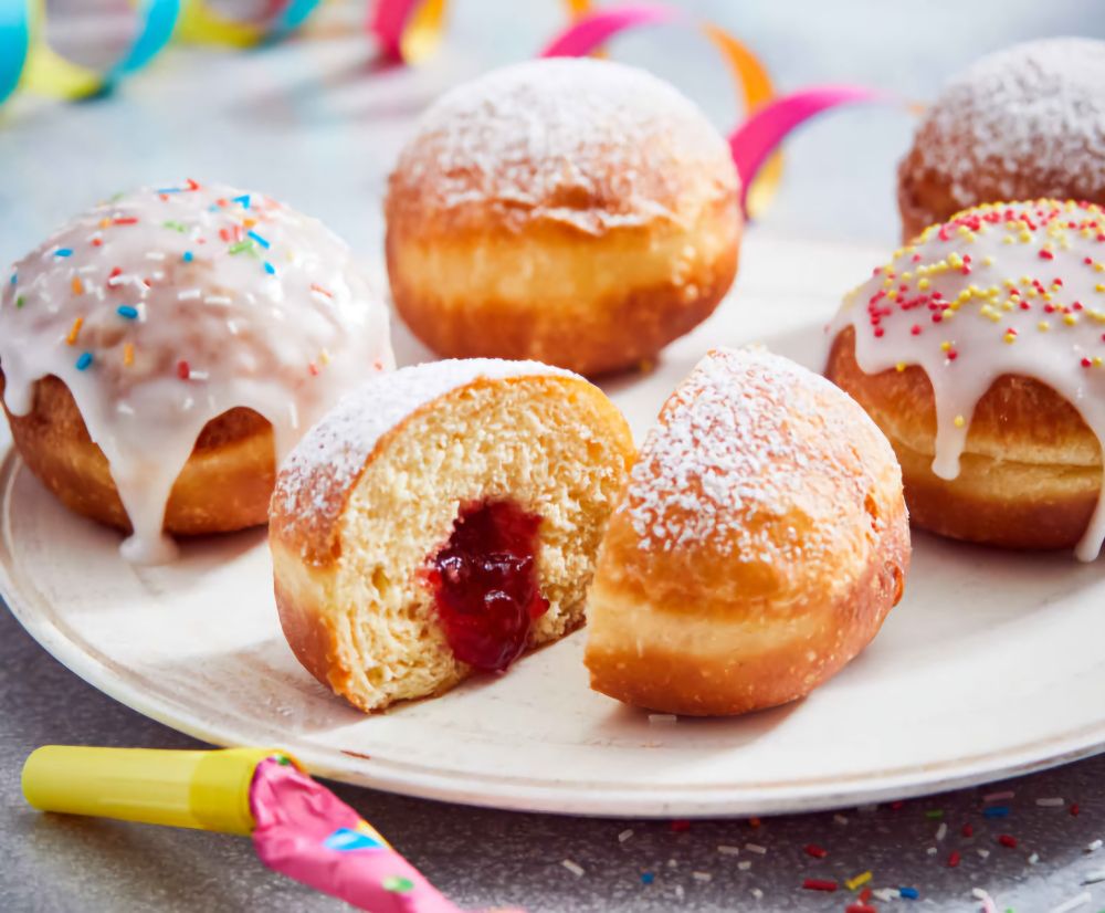 Mit roter Marmelade gefüllte Berliner, teilweise mit Zuckerguss überzogen, teilweise mit Zucker bestreut, auf einem weißen Teller.

Dahinter eine Luftschlange, davor eine Papierrollen-Tröte