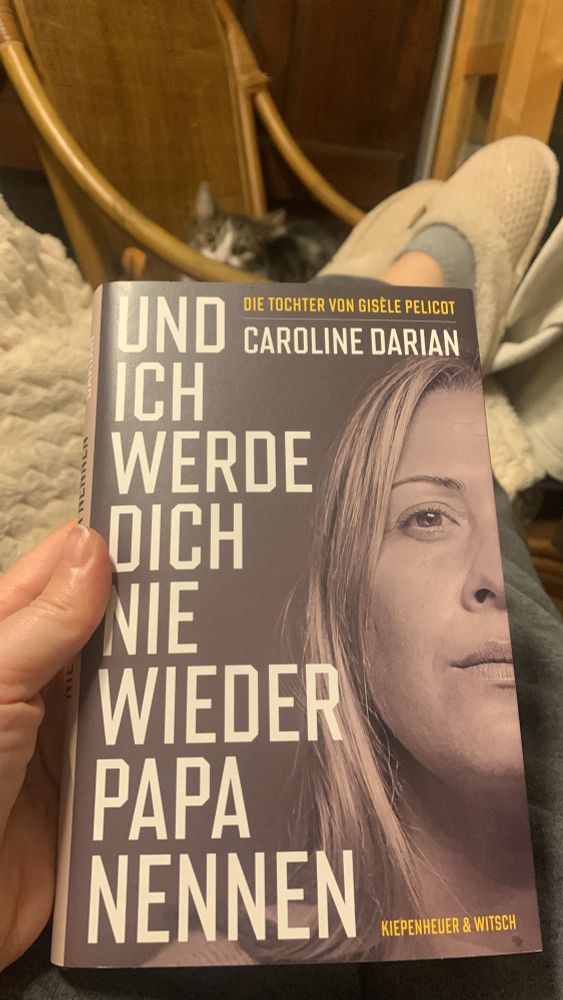 Meine Hand, die das Buch „Und ich werde dich nie wieder Papa nennen“ von Caroline Darian, der Tochter von Gisele Pelicot, hält. Im Hintergrund sieht man meine Katze im Schaukelstuhl 
