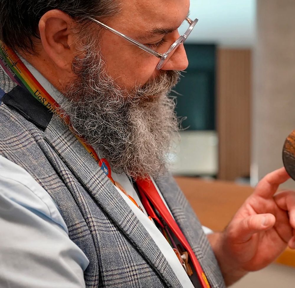 A man strokes the beak of a dodo replica, while wearing a red tie, rainbow-striped lanyard, dress shirt, and a plaid waistcoat with lapels. There’s a lot going on between his shoulder and his beard.