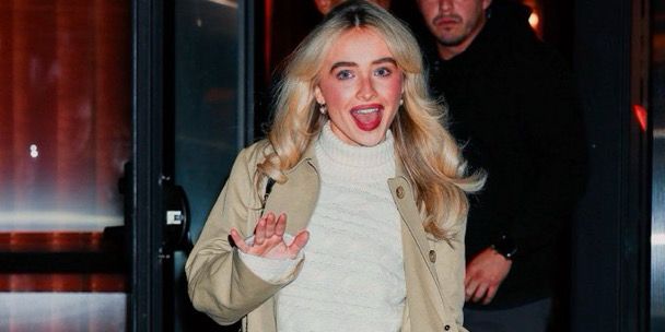 Sabrina Carpenter with long blonde hair wearing a beige coat and white sweater smiles and waves while exiting a building at night. A man in dark clothing stands behind her.