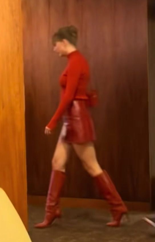 Taylor Swift wearing a red long-sleeve top, red mini skirt, and knee-high red boots is walking indoors near wooden walls, with her hair tied up.