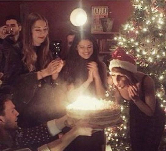 A group of people celebrate around a birthday cake with lit candles. Taylor Swift in a Santa hat leans forward to blow out the candles. A decorated Christmas tree glows in the background. Everyone looks happy.