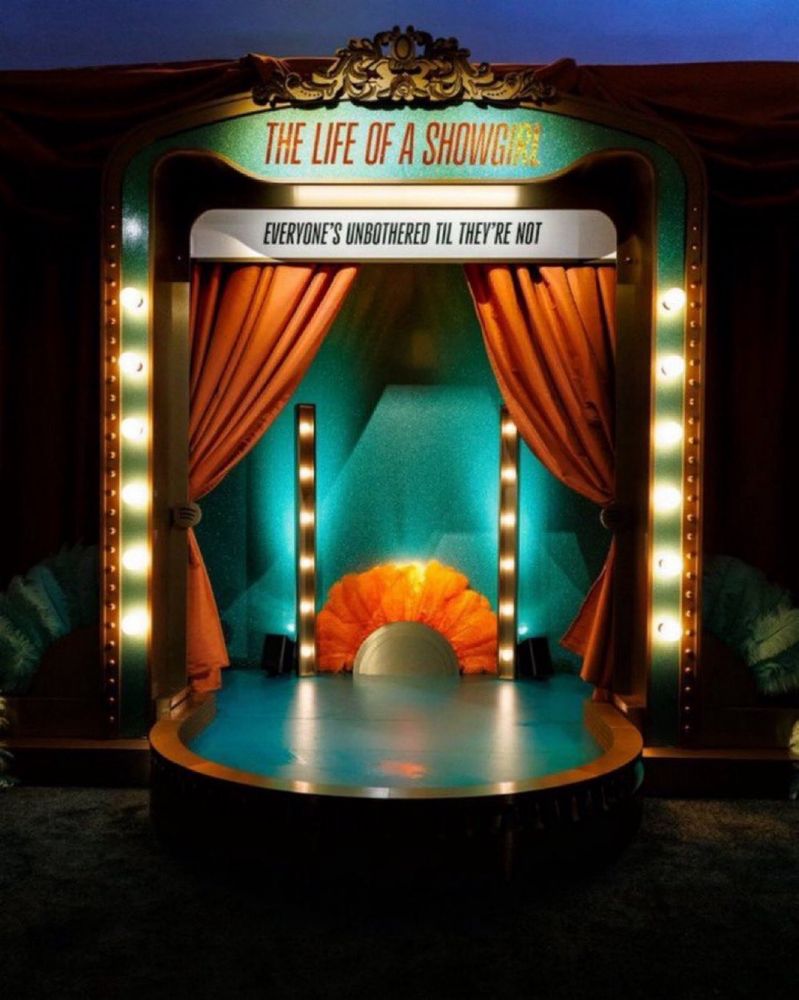 A glamorous, lit stage display with orange curtains and a sign reading THE LIFE OF A SHOWGIRL. Another sign says, EVERYONE’S UNBOTHERED TIL THEY’RE NOT. Feathers and lights add to the theatrical atmosphere.
