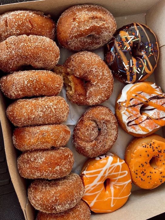 A box filled with cinnamon sugar donuts on the left and assorted decorated donuts with chocolate and orange icing on the right, some topped with sprinkles and drizzled icing.