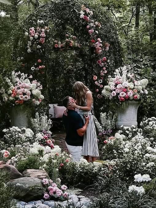 Travis down on one knee while taylor holds his head in her hands. They’re surrounded by flowers.