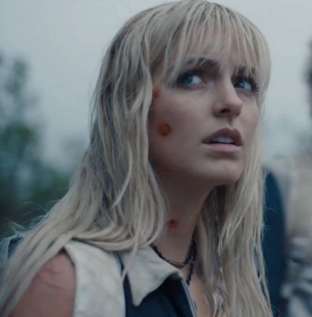 Maysilee Donner played by McKenna Grace in the hunger games: sunrise on the reaping movie with long, wet blonde hair and visible wounds on her face and neck looks up with a concerned expression. The background is blurry, suggesting an outdoor setting.