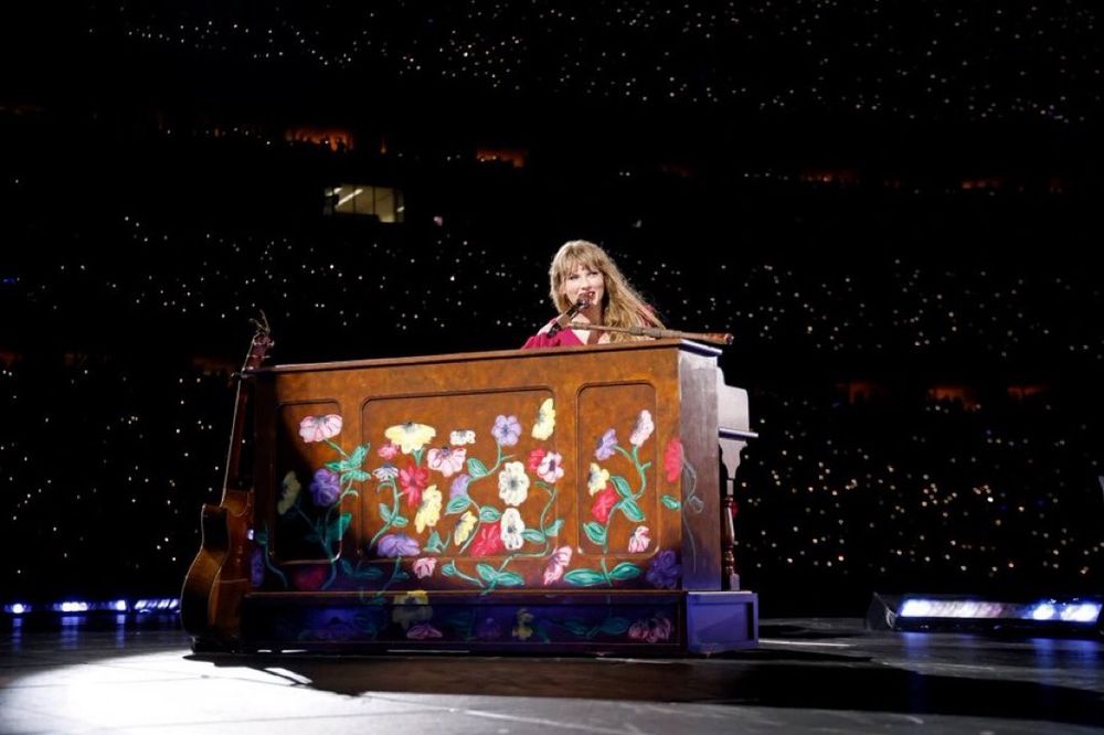 Taylor Swift plays a piano painted with colorful flowers on a dark stage, surrounded by an audience with small lights in the background.
