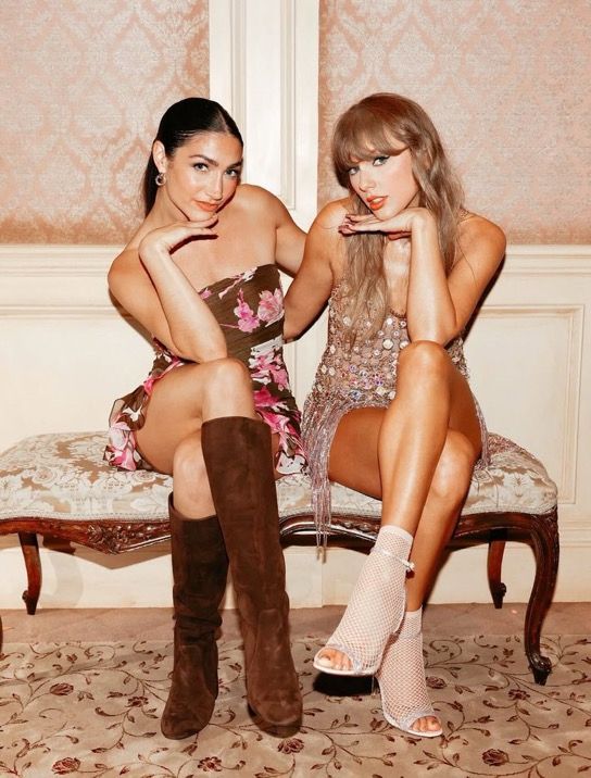Sydney Moss and Taylor Swift sit on an ornate bench, posing with one hand under their chins. Sydney Moss is wearing a floral dress with brown boots, Taylor Swift is wearing a sparkly dress with white mesh heels. They smile in a softly lit, elegant room with patterned wallpaper.