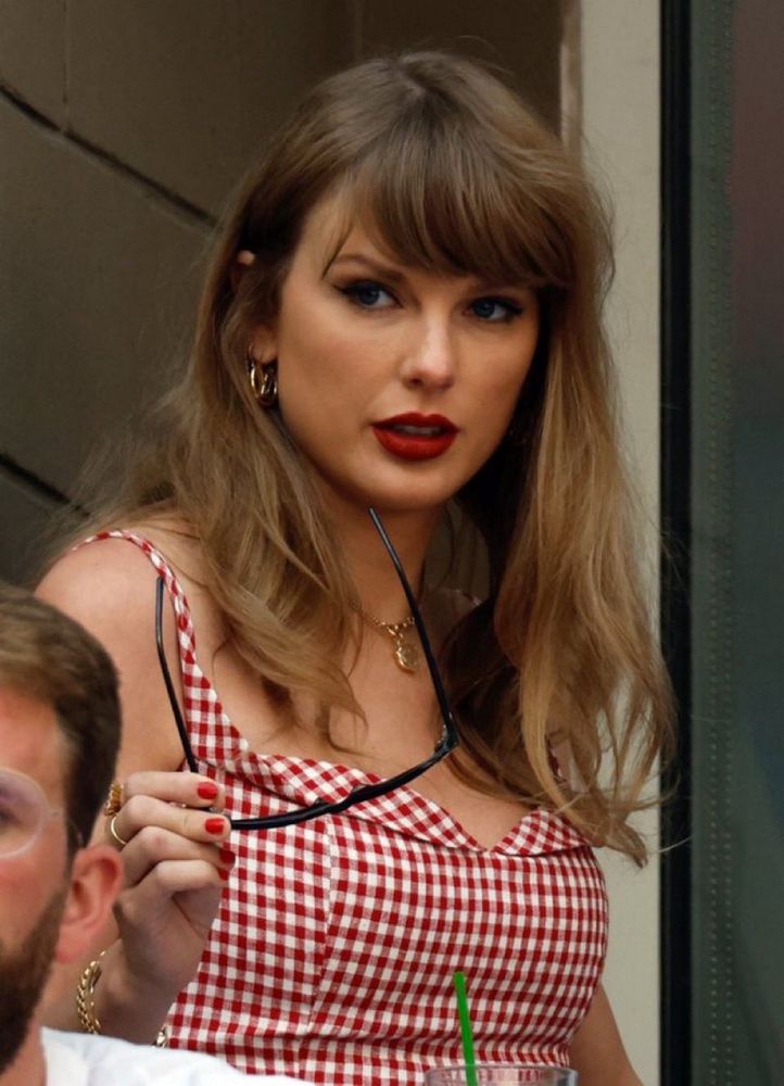 Taylor Swift with long blonde hair, red lipstick, and a red and white gingham dress wearing a gold heart necklace. 
