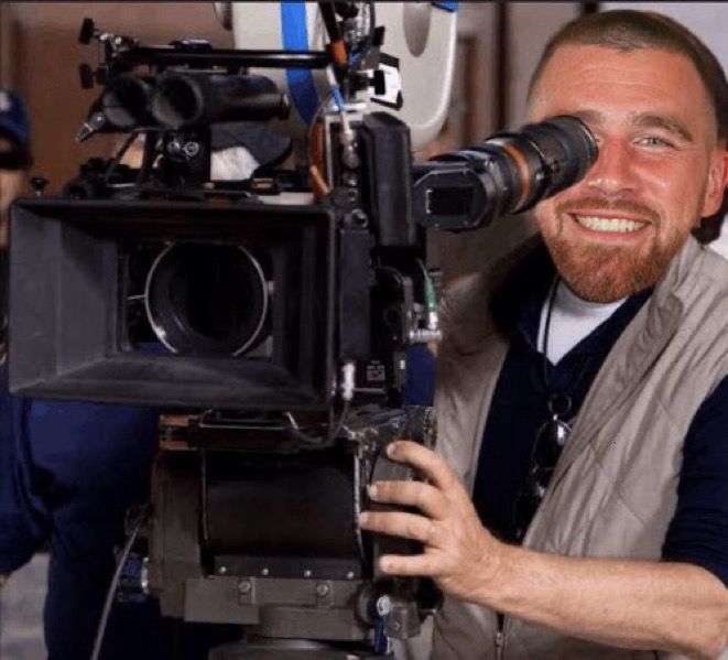A meme with Travis Kelce in a light-tan vest operates a large, professional movie camera.