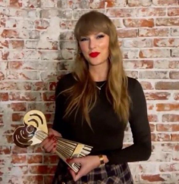 Taylor Swift, holding her iHeart award for tour of the century, smiles in front of a brick wall. Wearing a black long sleeve top and a plaid skirt, with red lipstick and red nails. 
