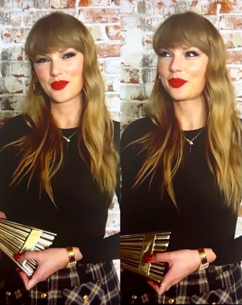 Taylor Swift in a black top, plaid skirt, red lips and
red nails and a gold diamond encrusted watch on with her iHeart award for tour of the century posing in front of a brick wall.