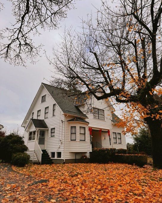 A white two-story house stands surrounded by trees with orange autumn leaves. Fallen leaves cover the ground, and the sky is overcast, creating a cozy autumn atmosphere. Red bows decorate the front porch.

