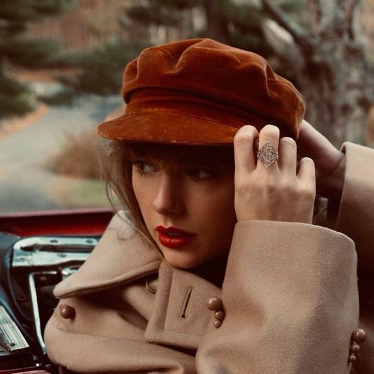 Taylor Swift for her red TV album cover in a tan coat and brown cap sits in a classic car, touching her hat. She has wavy hair, red lipstick, and wears a large ring. The background features trees and a blurred roadway.