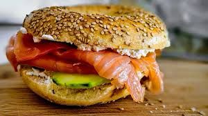 A sesame seed bagel sandwich filled with smoked salmon, cream cheese, and cucumber slices sits on a wooden surface.