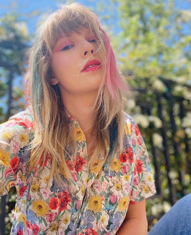 Taylor Swift with long, blonde hair with pink and blue streaks poses outdoors, wearing a floral shirt. They have bright pink eye makeup and lipstick. Green foliage and a fence are visible in the sunlit background.