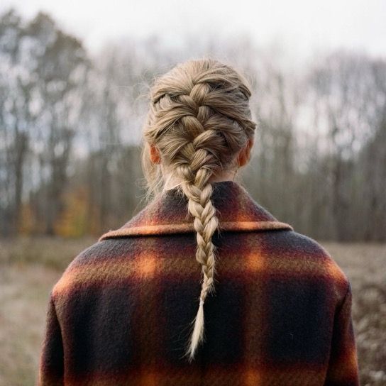 Taylor Swift for her evermore album cover with blonde hair in a French braid stands outdoors, facing away. They are wearing a plaid coat, and blurred trees are visible in the background under an overcast sky.