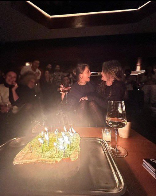 Taylor Swift and Selena Gomez sit close together at a dimly lit table, smiling and holding drinks, with a birthday cake topped with lit candles in the foreground and a group of people sitting and watching in the background.