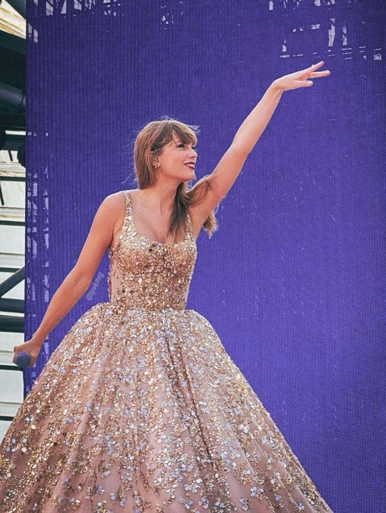 Taylor Swift in a sparkling gold dress stands on stage at the eras tour in front of a purple backdrop, holding a microphone and raising her arm with a joyful expression.