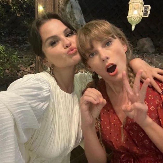 Taylor Swift and Selena Gomez pose closely for a selfie. Selena is wearing a white dress and makes a kissy face, Taylor Swift is in a red dress with braids holds up her hand and shapes her mouth in surprise. Both appear happy and playful.