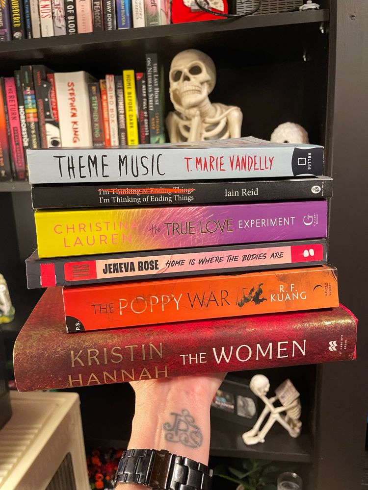 Stack of books. From top to bottom: Theme Music by T. Marie Vandelly, I’m Thinking of Ending Things by Iain Reid, The True Love Experiment by Christina Lauren, Home is Where the Bodies Are by Jeneva Rose, The Poppy War by R. F. Kuang and The Women by Kristin Hannah 