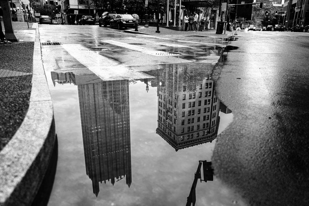 A black and white street scene in downtown Pittsburgh. There is a large puddle which is reflecting two skyscrapers. 