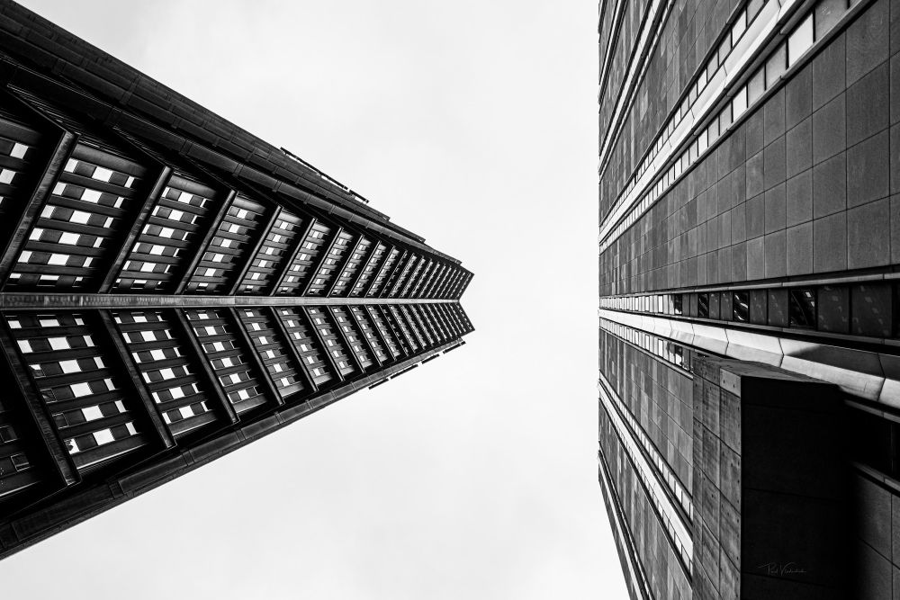 A photograph viewed straight up from the ground. On the left, the structure of the US Steel tower and on the entire right side of the image the flat wall of the Koppers Tower. The sky is entirely featureless white. 