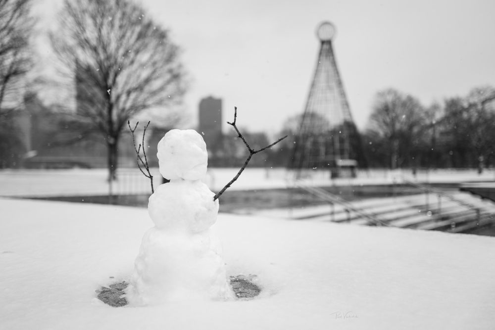 A photo of a snowman at Point State park in Pittsburgh. Blurred in the background is the Duquense Light Christmas tree. The city is visible in the background 