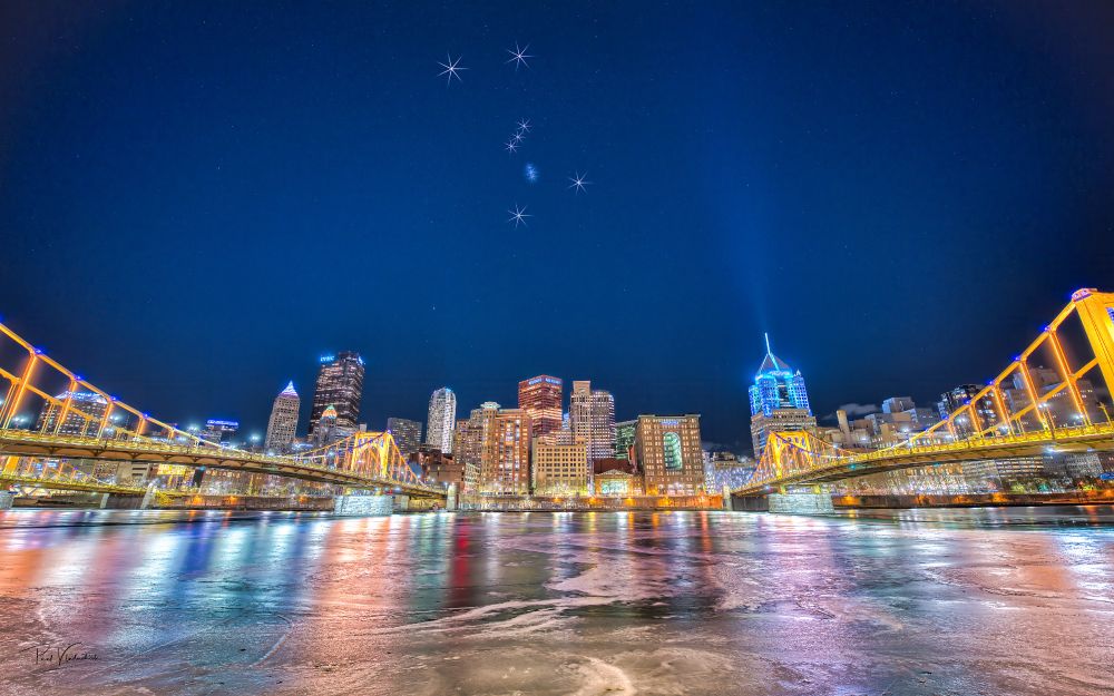 A photograph of Pittsburgh famed on either side by two of the three "Three Sisters bridges". The river is frozen. Orion has been highlighted by starbursts on each of the starts in the sky