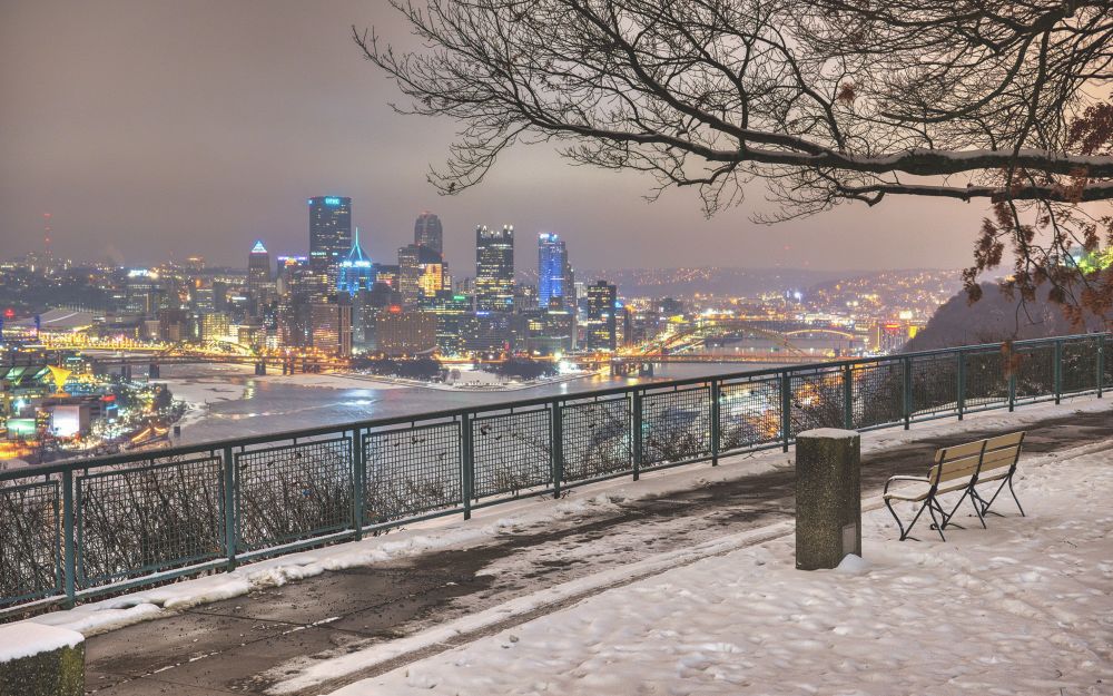 Photograph of downtown Pittsburgh from the West End Overlook. In the foreground is a snow covered lawn with a park bench and it in the background and slightly out of focus is the skyline of the city. 