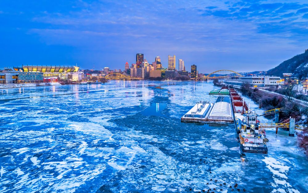 A photograph of Pittsburgh from the West End Bridge. The Ohio River is frozen over. A Barge is iced in on the bank of the river. 