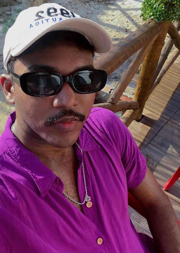 selfie sentado, usando camisa lilás de botão, correntinha de aço no pescoço, óculos escuros e boné bege.